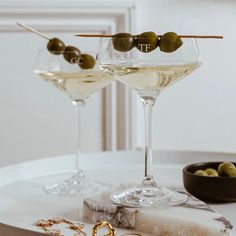 two wine glasses filled with white wine and olives on a table next to a pair of gold hoop earrings