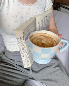 a woman sitting on a bed holding a cup of coffee and an open book in her lap