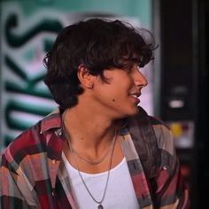 a young man with black hair wearing a checkered shirt and silver necklace, standing in front of a vending machine