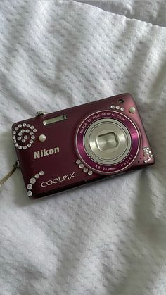 a red digital camera sitting on top of a bed next to a white sheet with the word nikon printed on it