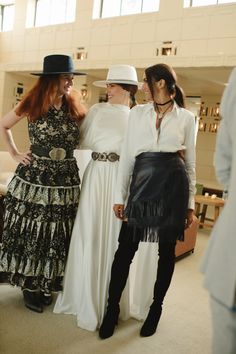 three women standing next to each other in front of a white wall wearing dresses and hats