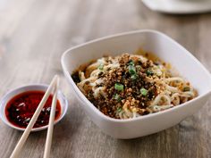 a bowl of noodles and chopsticks on a wooden table next to some dipping sauce