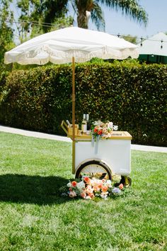 an umbrella is on top of a cart with flowers and bottles in the grass near a hedge