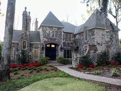 a large stone house surrounded by trees and flowers in the front yard, with a walkway leading up to it