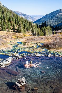 two people in a hot tub surrounded by rocks and water with mountains in the background