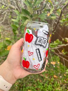 a hand holding up a glass with an apple and book design on the bottom, in front of some trees