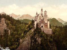 an old castle sitting on top of a cliff in the middle of a forest with mountains behind it