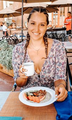 a woman sitting at an outdoor table with a plate of food and cup of coffee