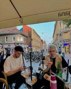 two people sitting at an outdoor table with wine glasses in front of them on the street