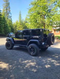 a black jeep parked in the middle of a parking lot with trees and bushes behind it