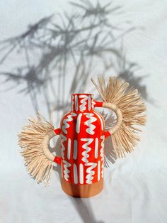 a red vase sitting on top of a white table next to a plant and shadow