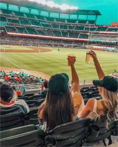 two women are sitting in the stands at a baseball game and one is holding up a cup