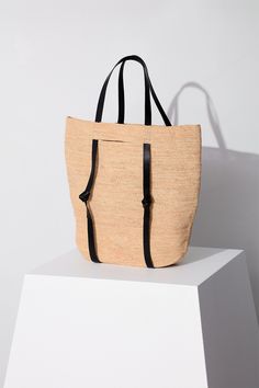 Design Tanner is a signature beach bag, meticulously crafted from durable raffia straw, accented with regenerative black leather details, and featuring a straps for a practical hat holding feature. Material Our raffia is responsibly sourced from a vertically-integrated farm in Madagascar where farmers hand-harvest fibers from the leaves of the native raffia palm, then dry and braid them. Each piece is naturally resilient to water and sun, allowing it to harmonize with natural environments. At th Modern Travel Straw Bag, Modern Woven Leather Beach Bag, Modern Bucket Bag With Braided Handles For Beach, Modern Beach Bucket Bag, Modern Natural Bucket Bag For Summer, Modern Handwoven Natural Straw Bag, Modern Woven Natural Straw Bag, Modern Summer Beach Bucket Bag, Modern Bucket Bag With Braided Handles For Vacation