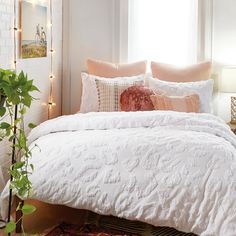 a bed with white comforter and pillows next to a potted plant in front of a window