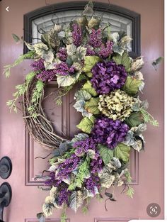 a wreath with purple flowers and green leaves hanging on a pink front door, next to a pair of black shoes