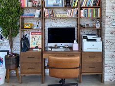 a desk with a computer, printer and bookshelf in front of a brick wall