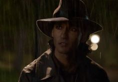 a man wearing a hat standing in the rain at night with his head turned to the side