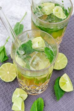 two glasses filled with limeade and mint on top of a purple cloth next to sliced limes