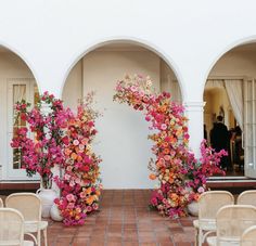 an archway decorated with pink and orange flowers in front of a white building filled with chairs