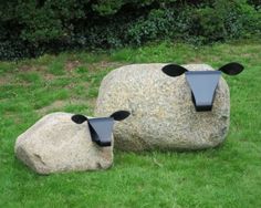 two black sheep sculptures sitting on top of a green field next to a large rock