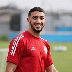 a man standing on top of a soccer field wearing a red shirt and smiling at the camera