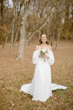 a woman in a white dress holding a bouquet and posing for the camera with trees in the background