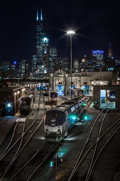 a train station with several trains on the tracks and city lights in the background at night