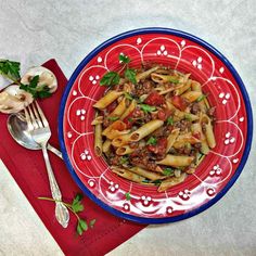 a red and blue plate topped with pasta next to a silver spoon on top of a red napkin