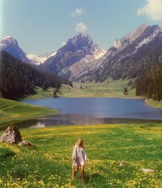 a woman standing on top of a lush green field next to a lake surrounded by mountains