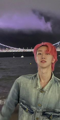 a man with pink hair standing in front of the water and a bridge at night