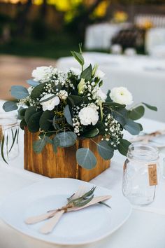 the table is set with white flowers and greenery in a wooden box on it