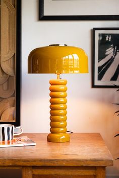 Lifestyle image of the Modern Metal Mustard Table Lamp illuminated and displayed on a wooden desk surrounded by various other home accessories. Squashy Sofas, Faculty Office, Bee Cafe, Unusual Table Lamps, Quirky Table Lamp, Quirky Table, Checkerboard Table, Yellow Table Lamp, Yellow Lamp