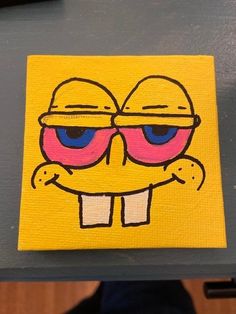 a yellow square with blue eyes and pink glasses on it, sitting on a table