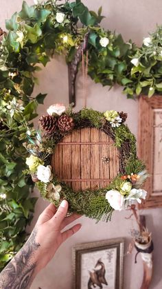 a person holding up a wreath with pine cones and white flowers on the front, surrounded by greenery