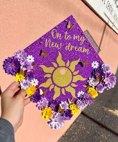 a hand holding up a purple and yellow graduation cap with flowers on it that says, on to my very dream