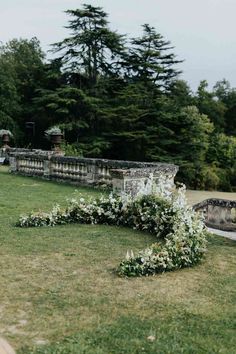 a wedding arch with white flowers on it in the middle of a grassy area next to a stone wall