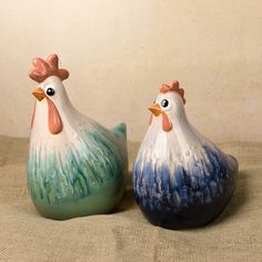 two ceramic chickens sitting next to each other on top of a cloth covered tablecloth