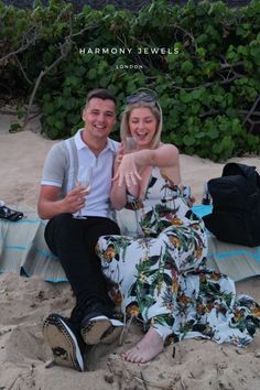 a man and woman sitting on top of a sandy beach next to each other holding hands