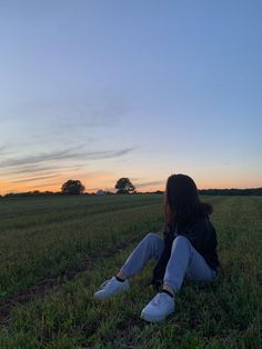 a woman sitting on the ground in a field at sunset with her back to the camera