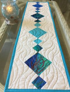 a quilted table runner with blue and green designs on the edge, sitting on a glass table