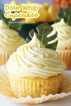 two cupcakes with white frosting on top and green leaves in the background