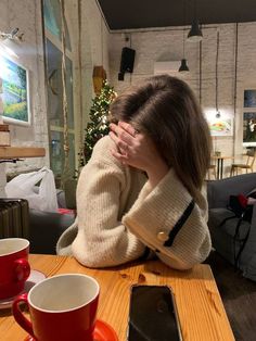 a woman covering her face while sitting at a table with two cups and a cell phone