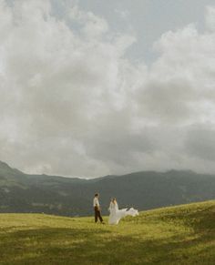 a bride and groom are walking in the grass with mountains in the backgroud