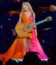 a woman in an orange and pink dress holding a guitar while standing on top of a stage