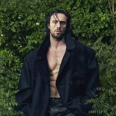 a shirtless man standing in front of some bushes and trees with his hands in his pockets