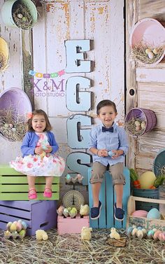 two young children sitting on crates in front of an easter backdrop with eggs and hay