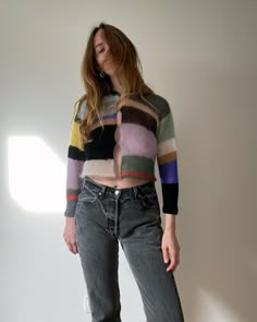 a woman standing in front of a white wall with her hands on her hips, wearing jeans and a multicolored sweater