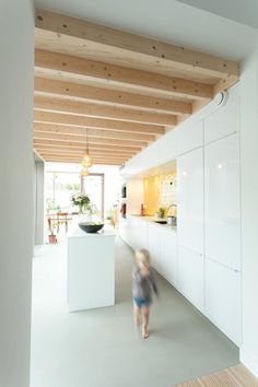 a woman walking through a kitchen next to a white counter top and wooden flooring