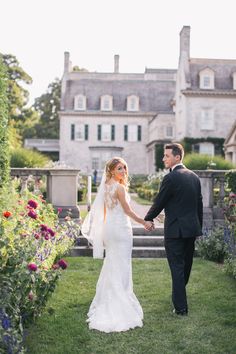 a man and woman are holding hands in front of a large house with flowers on the lawn