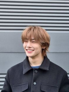 a young man standing in front of a wall wearing a black coat and smiling at the camera